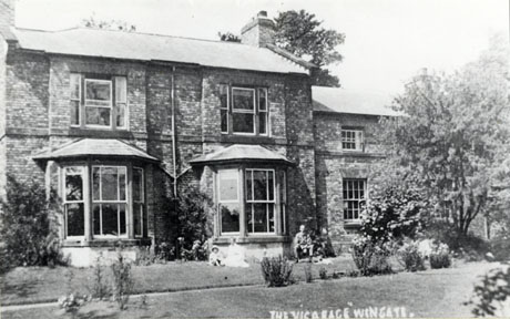 Photograph of the exterior of a large brick house with two large bay windows on the ground floor and two large windows on the first floor; on the right of the picture is an extension to the house partly obscured by trees; a woman and a child are sitting on the grass on the left of the right-hand bay window and a man is sitting on a seat on the other side of that bay; they are too indistinct to be recognised; in the foreground of the photograph are lawns and bushes; the photograph has a caption reading The Vicarage, Wingate