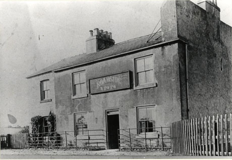Photograph of the facade of a building of brick with stucco on it; it has six windows and a doorway in the centre; in front of the building is an iron fence and the road; a fence of wood curves round at the right of the picture; above the door of the building is a sign reading Roadside Inn; the inn has been identified as being in Wingate