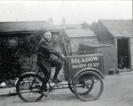 Photograph showing a boy aged approximately fourteen years sitting on a tricycle with a container at the front with the words Meadow Dairy Co. Ltd. on it; behind the boy and tricycle are bushes and a shed with the roofs of houses; the boy has been identified as being in Wingate