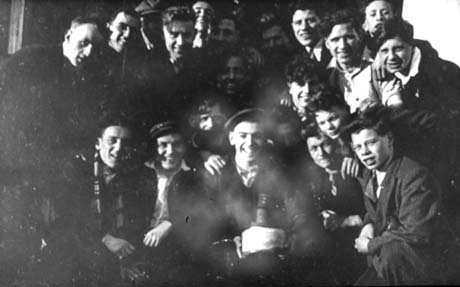 Photograph showing the faces of a group of sixteen men laughing and wearing suits and scarves; it is not possible to determine the occasion for the group's coming together, but the man at the front is holding what may be a cake in his hands; they have been identified as being in Wingate