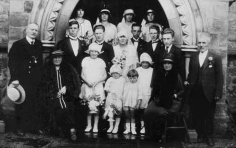 Photograph of a newly married couple outside the west door of a church; they are accompanied by four child bridesmaids, aged between three and ten years, standing in front of the bride and groom; behind the bride and groom are five young men and, behind them, four young women, possibly bridesmaids; on either side of the bride and groom are a middle aged man and woman, the latter of whom are seated; the bride has been identified as Eva, daughter of Jack Ord and the wedding has been identified as taking place in Wingate