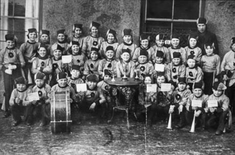 Photograph of thirty four children, aged approximately between ten and fifteen years, in uniform and posed against the wall of a building; eight of them are displaying cards and there is a table in front of the group on which there are three trophy cups; a drum is in front of one child on the front row; a man is standing at the back of the group on the right-hand side; the group has been identified as a jazz band in Wingate