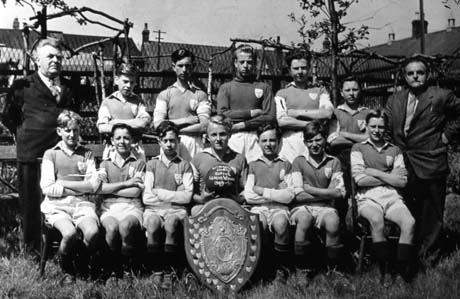Photograph of twelve boys, aged approximately between twelve and fifteen years, in football strip, posed in a garden; they are accompanied by two men; a large shield is propped in front of the group; a boy on the front row is holding a football on which is written: Wingate County School League Winners 1949-50