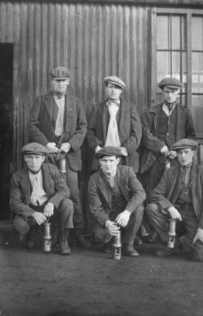 Photograph of six men grouped in front of a corrugated iron building; all of them are dressed in caps, waistcoats, jackets, scarves and trousers and are carrying miner's lamps; they have been identified as a Group of Miners, Wingate