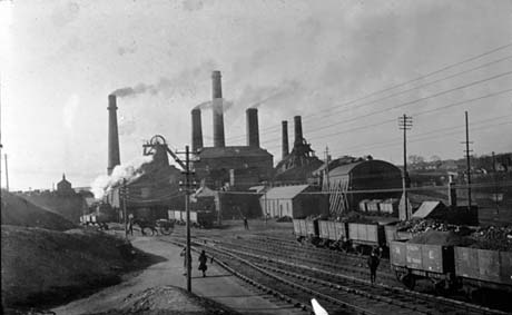 Photograph showing heaps of waste on the left, and rails running away from the camera in the middle; seven loaded coal waggons are on the rails; eight waggons are on the rails in the distance and a horse and cart can be seen crossing the rails at the left; beyond the rails are the buildings, winding gear and six chimneys of a colliery, which has been identified as Wingate Colliery