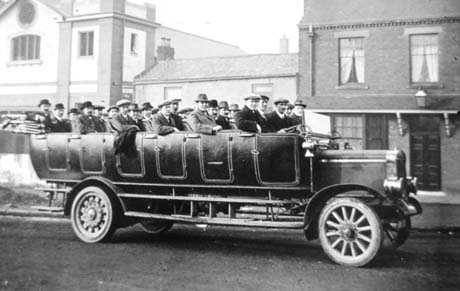 Photograph showing the side of a motor charabanc with approximately thirty men sitting in it; behind the charabanc are an imposing building and two houses; the men are described as being about to set out on an outing from Wingate