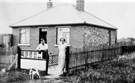 Photograph showing a man and a woman standing at the gate of a bungalow with leaded windows; the woman is dressed in a frock and the man in shirt sleeves; a terrier dog is standing with them in front of the gate; they have been identified as Mr. and Mrs. Billy Howe of Wingate at their Bungalow at Crimdon