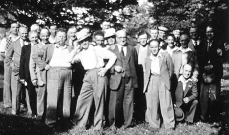 Photograph of approximately twenty five men standing under trees; they are dressed in suits and open shirts and in ties and suits; they have been identified as Gardeners' Society, Wingate