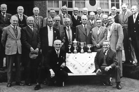 Photograph of twenty three middle aged men grouped against the window of a building; all are dressed in suits and ties; in front of the group is a table on which there are four trophy cups; the photograph has been identified as Bowls Club Presentation, Wingate