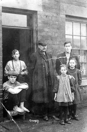 Photograph showing a young man, in shirt sleeves with bare feet and wearing a cap, sitting on the doorstep of a terraced house and resting his feet on a stool; next to him is another young man standing in the doorway of the house in shirt sleeves; next to him is a middle-aged man wearing a cap, overcoat, suit and waistcoat leaning against the wall of the house near the doorway; on the right are a young man wearing a jacket with two girls, aged approximately eight and nine years, standing in front of him; the girls are wearing dark stockings a pinafore and a dress; they have been identified as First Miner's Family, Wingate