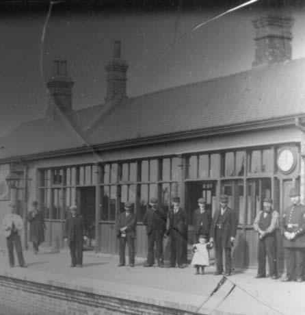Photograph showing a low building with extensive windows and a clock on it with a railway platform in front of it, presumably the waiting room of a railway station; in front of it are six men dressed in coats and suits; in addition, there are three men dressed in the uniform of the railway company and a policeman; one of the employees of the railway company is holding the hand of a small girl, aged approximately three years; the photograph has been identified as Wellfield Station, Wingate