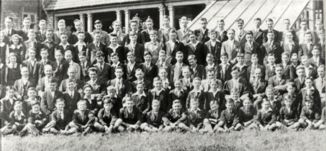 Photograph showing a hundred and five boys and masters, posed outside buildings identified as those of the A. J. Dawson Grammar School, Wingate; the photograph adjoins wing0135 on the right and wing0137 on the left